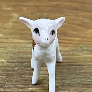 May include: A small white ceramic figurine of a lamb with black eyes and a pink nose. The lamb is standing on all four legs and has a brown and white patterned design on its back.