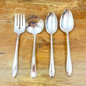 May include: A set of four silver-toned metal utensils labeled SET 1, including a three-tined serving fork, a round-bowled ladle, a small spoon, and a large oval-bowled spoon, all featuring decorative embossed patterns on the handles.