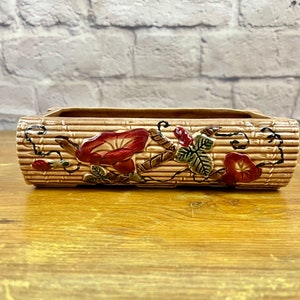 May include: Rectangular ceramic planter with a tan, ribbed exterior. The planter is decorated with red and brown morning glory flowers and green leaves. The planter sits on a wooden surface.