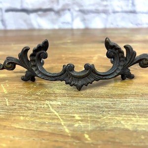 Puede incluir: Tirador de cajón de bronce adornado con un diseño de volutas decorativas. El tirador mide aproximadamente 10 cm de largo y tiene una pátina oscura.