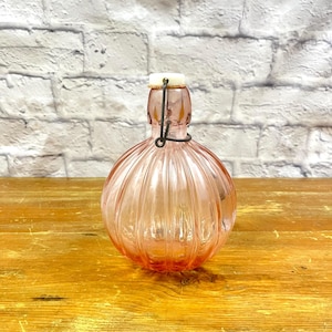 May include: A pink glass bottle with a ribbed design. The bottle has a white stopper and a wire closure. The bottle is sitting on a wooden surface.