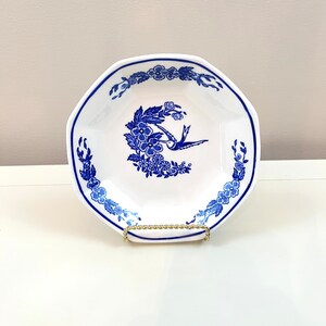 以下が含まれることがあります： 青い花柄と青い鳥が描かれた白い八角形のセラミックボウル。ボウルは金色のスタンドの上に置かれています。