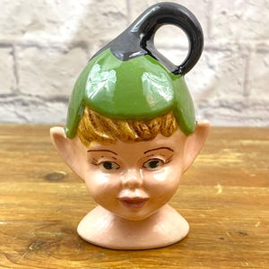 May include: A ceramic elf head figurine with a green hat and black handle. The elf has tan hair, large ears, and a smiling face with blue eyes. The figurine is on a wooden surface.