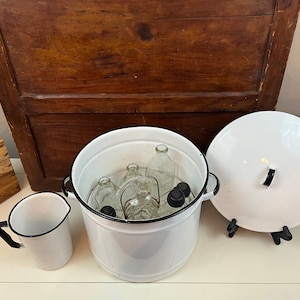 May include: White enamelware container with a black rim and lid. The container is filled with glass bottles and jars with black lids. The container is sitting on a white surface.