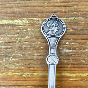 May include: Silver spoon with a round, ornate handle featuring a profile of a person wearing a helmet. The handle is decorated with a floral design.