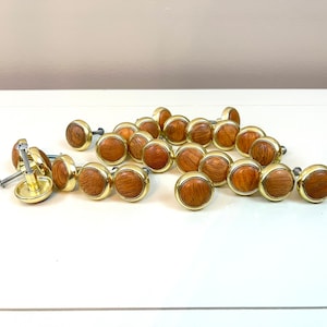 Op de afbeelding: Een verzameling ronde houten kastknoppen met een goudkleurige metalen rand. De knoppen hebben een natuurlijk houtnerfpatroon en zijn gerangschikt op een wit oppervlak. Sommige knoppen hebben schroeven.