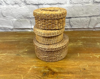 May include: Three round, woven baskets stacked. The baskets are made of light brown material and have lids. The background is a white brick wall and a wooden surface. The baskets are likely for storage.