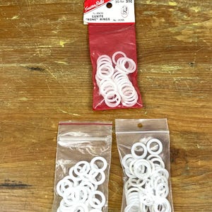 Op de afbeelding: Witte plastic ringen in doorzichtige plastic zakken en een rode plastic zak. De rode zak heeft een label met "Susan Bates" en "5/8 inch Luxite 'Bone' Rings". Op een houten oppervlak.