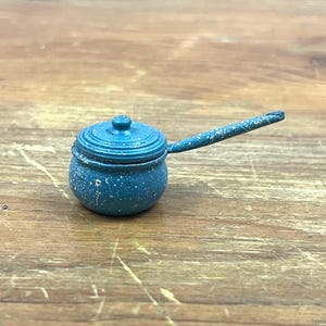 May include: A small, blue toy saucepan with a lid and a long handle. The saucepan is speckled with white paint and sits on a wooden surface. The handle is approximately 4 inches long.