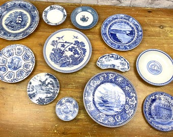 May include: A collection of blue and white decorative plates with various designs, including ships, landscapes, and architectural scenes. The plates are arranged on a wooden surface, showcasing a vintage aesthetic.