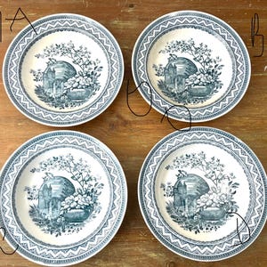 May include: Four white ceramic plates with a blue floral and architectural design. Each plate features a central image of a building and flowers, surrounded by a detailed blue border. The plates are arranged on a wooden surface.