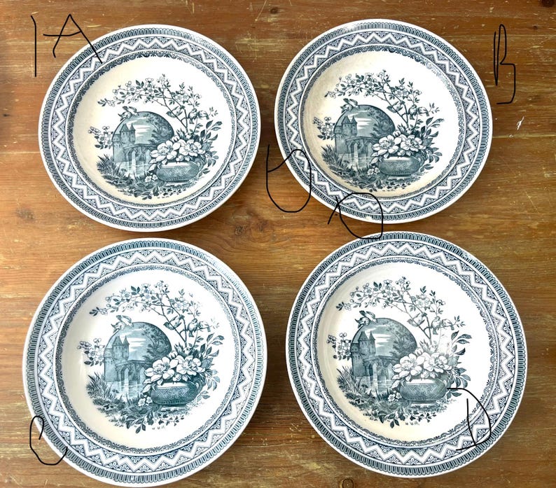 May include: Four white ceramic plates with a blue floral and architectural design. Each plate features a central image of a building and flowers, surrounded by a detailed blue border. The plates are arranged on a wooden surface.
