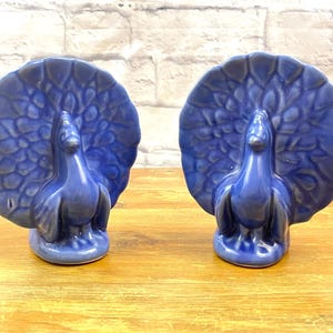 Könnte beinhalten: Zwei kobaltblaue Keramik-Pfauenfiguren. Jeder Pfau hat einen detaillierten, fächerförmigen Schwanz und steht aufrecht. Die Figuren werden auf einer Holzoberfläche vor einer weißen Backsteinmauer ausgestellt.