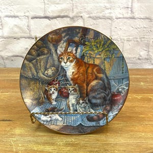 May include: A round decorative ceramic plate featuring a detailed illustration of a large ginger tabby cat sitting with three small kittens on a blue patterned cloth. The background shows a burlap sack of potatoes and a wooden basket of green beans.