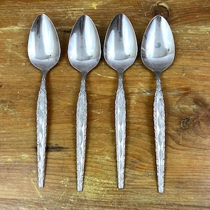 May include: Four silver spoons with oval bowls and ornate, textured handles are arranged on a wooden surface. The spoons are reflective, and the handles feature a repeating pattern. The spoons are a set, likely for dining.