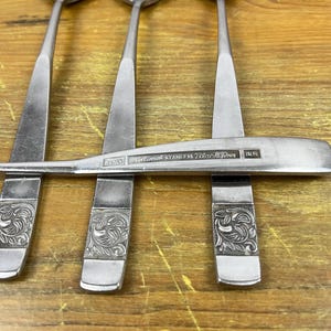 May include: Close-up of stainless steel spoons with a floral design on the handles. The spoons are arranged on a wooden surface. The text "National Stainless Black Rose" is visible on one of the spoons.