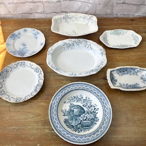 May include: A selection of antique white and blue porcelain dishes, including plates and serving platters, displayed on a wooden surface. The dishes feature floral and botanical patterns, with a vintage aesthetic.