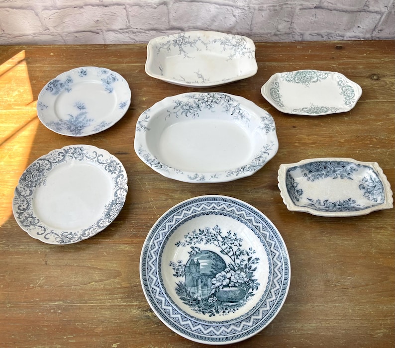 May include: A selection of antique white and blue porcelain dishes, including plates and serving platters, displayed on a wooden surface. The dishes feature floral and botanical patterns, with a vintage aesthetic.