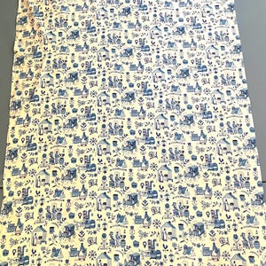 以下が含まれることがあります： 青とクリーム色の繰り返し模様の布。小さな家、馬車、人々、花の要素がデザインされています。全体的な美しさは、ヴィンテージのトワル生地を彷彿とさせます。