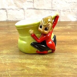 Puede incluir: Una figurita de cerámica de un elfo con un atuendo rojo y negro, sujetando una maceta verde en forma de bota. El elfo tiene una expresión caprichosa y ojos grandes.