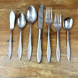 May include: A set of seven stainless steel flatware pieces including a butter knife, two spoons, a dinner knife, two forks, and a small spoon. Each handle features a raised, repeating leaf-like pattern along the center, with a polished finish.