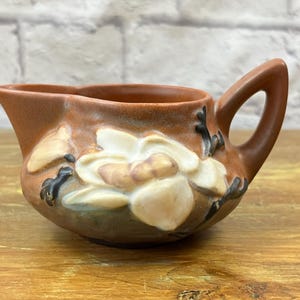 Puede incluir: Una cremaera de cerámica marrón con asa y boquilla. La cremaera está decorada con un diseño floral en relieve en tonos blanco, beige y negro. La cremaera está sobre una superficie de madera.