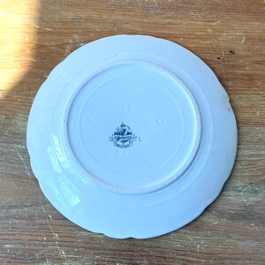 May include: A white ceramic plate with a scalloped edge, seen from the underside. The plate features a central circular area and a decorative emblem in the centre. It rests on a wooden surface.