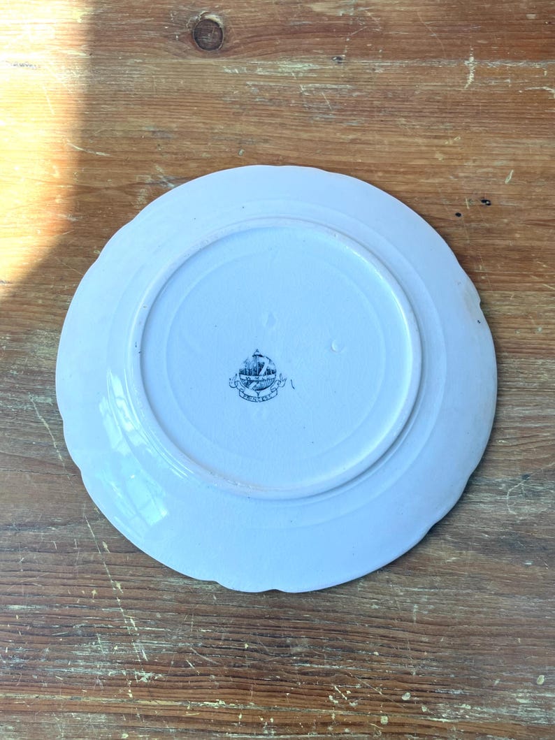 May include: A white ceramic plate with a scalloped edge, seen from the underside. The plate features a central circular area and a decorative emblem in the centre. It rests on a wooden surface.