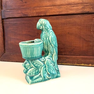 May include: A teal green ceramic parrot planter with a small pot on its perch. The parrot is perched on a branch with leaves. The planter is marked "USA" on the bottom.