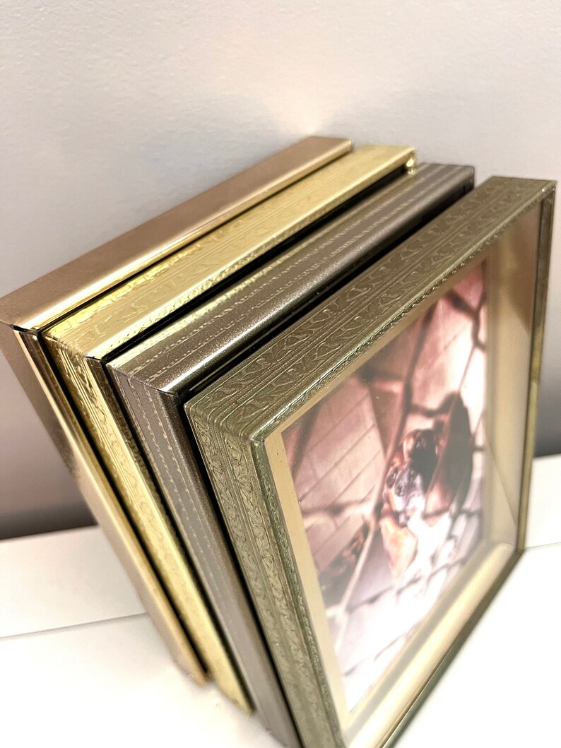 May include: Four picture frames stacked on top of each other. The top frame is a dark brown color, the second frame is a gold color, the third frame is a light brown color, and the bottom frame is a green color. All frames have an ornate design around the edges.