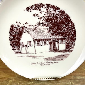 May include: A white ceramic plate with a brown line around the edge. The plate features a brown line drawing of a house with trees and the text "Upper Providence Meeting House 1828-1978".