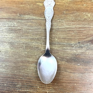 May include: A silver spoon with a detailed handle featuring an embossed design. The spoon's bowl is oval-shaped and reflects the light. The handle has a decorative top with a detailed scene. The spoon is set against a wooden background.