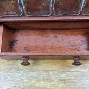 May include: A vintage wooden spice rack with a pull-out drawer and four glass jars. The rack is made of dark wood and has a warm, reddish-brown hue. The drawer has two small, round knobs. The glass jars have a decorative leaf design.