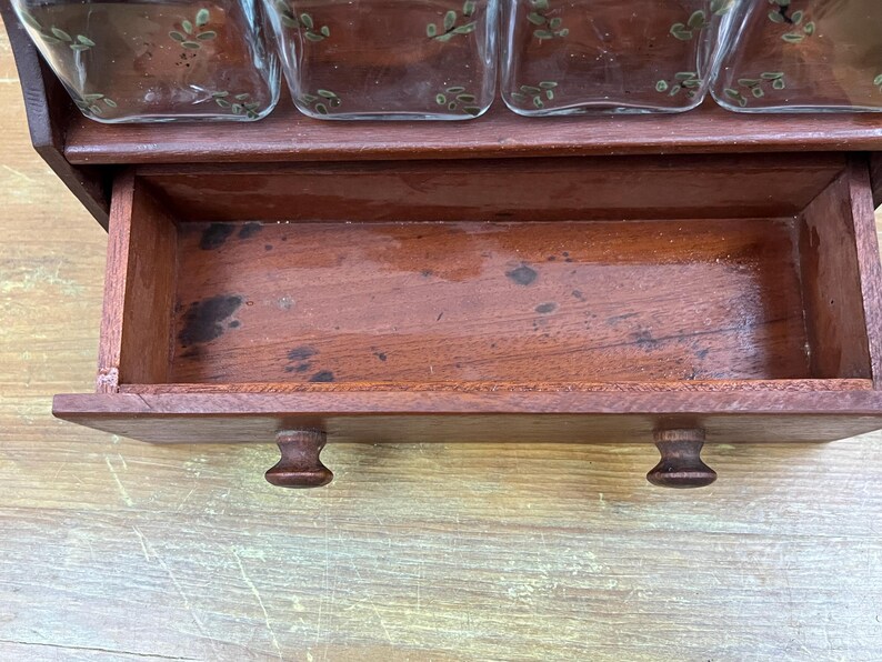 May include: A vintage wooden spice rack with a pull-out drawer and four glass jars. The rack is made of dark wood and has a warm, reddish-brown hue. The drawer has two small, round knobs. The glass jars have a decorative leaf design.