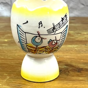 Può includere: Un portauovo in ceramica con un design stravagante. La tazza è bianca con bordo e base gialli. Presenta una scena dipinta di note musicali e due uccelli dei cartoni animati che suonano strumenti. Il design include accenti blu, rosa e gialli.