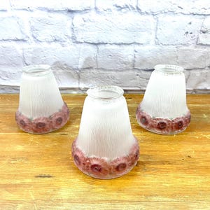 May include: Three white frosted glass lampshades with pink floral designs around the bottom edge. The lampshades are sitting on a wooden surface.