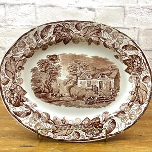 May include: An oval ceramic platter featuring a brown and white transferware pattern. The center depicts a rural cottage landscape with trees, while the wide rim is decorated with a detailed border of intertwined hop vines and leaves.
