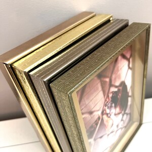 May include: Four picture frames stacked on top of each other. The top frame is a dark brown color, the second frame is a gold color, the third frame is a light brown color, and the bottom frame is a green color. All frames have an ornate design.