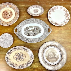 May include: A collection of seven vintage ceramic plates with brown and white floral patterns. The plates are arranged on a wooden surface.