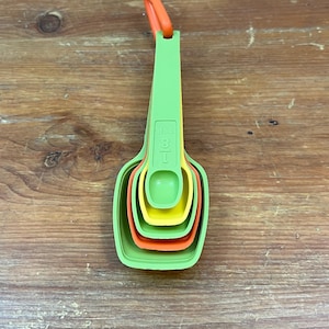 Può includere: Un set di quattro cucchiai dosatori colorati in verde, arancione e giallo. I cucchiai sono nidificati insieme e attaccati a un manico verde con un anello arancione. Sul manico è stampata la misura "1 TSP".