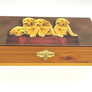 Könnte beinhalten: Rechteckige Holzkiste mit Scharnierdeckel. Der Deckel zeigt einen braunen Hintergrund mit Illustrationen von vier goldenen Labrador-Welpen. Die Box hat einen Messingverschluss und die Worte "New Hampshire" sind aufgedruckt.