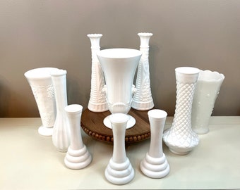 Puede incluir: Un conjunto de ocho jarrones de vidrio de leche blanco de varias formas y tamaños. Los jarrones están dispuestos sobre un pedestal de madera.