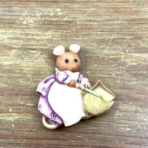 Könnte beinhalten: Eine Keramikfigur einer Maus in einem lila-weißen Kleid, die einen Besen hält. Die Maus hat braunes Fell und rosa Ohren. Die Figur steht vor einem hölzernen Hintergrund.
