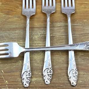 May include: Four silver forks with ornate floral designs on the handles. The forks are arranged in a criss-cross pattern.