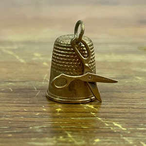 May include: A gold-toned thimble with a pair of small scissors attached to the top. The scissors are open and the thimble is sitting on a wooden surface.