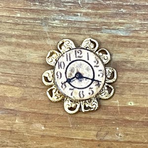 May include: A small, antique-style clock face with black numerals and hands, set within a gold-colored, flower-shaped frame. The clock face has a distressed, aged appearance, suggesting a vintage aesthetic. The hands point to approximately 9:18.