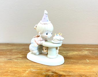 Może przedstawiać: Porcelanowa figurka dziecka w liliowym czapeczku urodzinowym w kropki, pochylonego nad małym okrągłym stolikiem, aby zdmuchnąć świeczki na torcie urodzinowym, podczas gdy mały szczeniak siedzi pod stołem na owalnej podstawie.