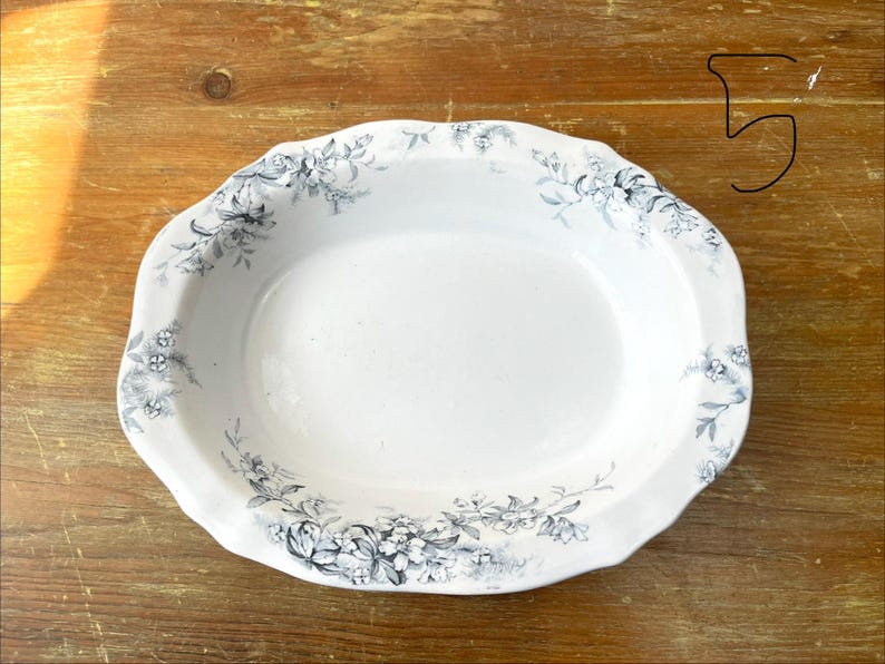 May include: Oval white serving dish with a scalloped edge, decorated with a floral pattern in shades of grey and blue. The dish has a smooth, glossy surface, likely made of ceramic or porcelain.