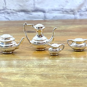 May include: A silver miniature tea set, including a teapot, creamer, sugar bowl, and a small pitcher. The set is displayed on a wooden surface. The teapot has a curved spout and a lid with a small knob.
