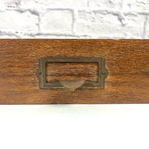 May include: A wooden drawer with a brass label holder. The drawer is a light brown color and has a wood grain pattern. The label holder is rectangular and has a slightly curved top.